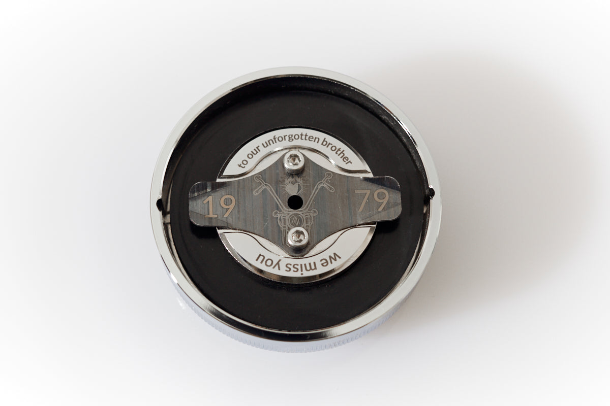 Close-up of the inside of a chrome screw-top lid from the Haus of Urns Freedom Urn, showing a personalised laser engraving on the inner plate. The engraving features a classic Frisco-style chopper motorcycle illustration flanked by the birth year 1979, with the inscriptions "to our unforgotten brother" and "we miss you" — a hidden memorial message visible only when the lid is removed.