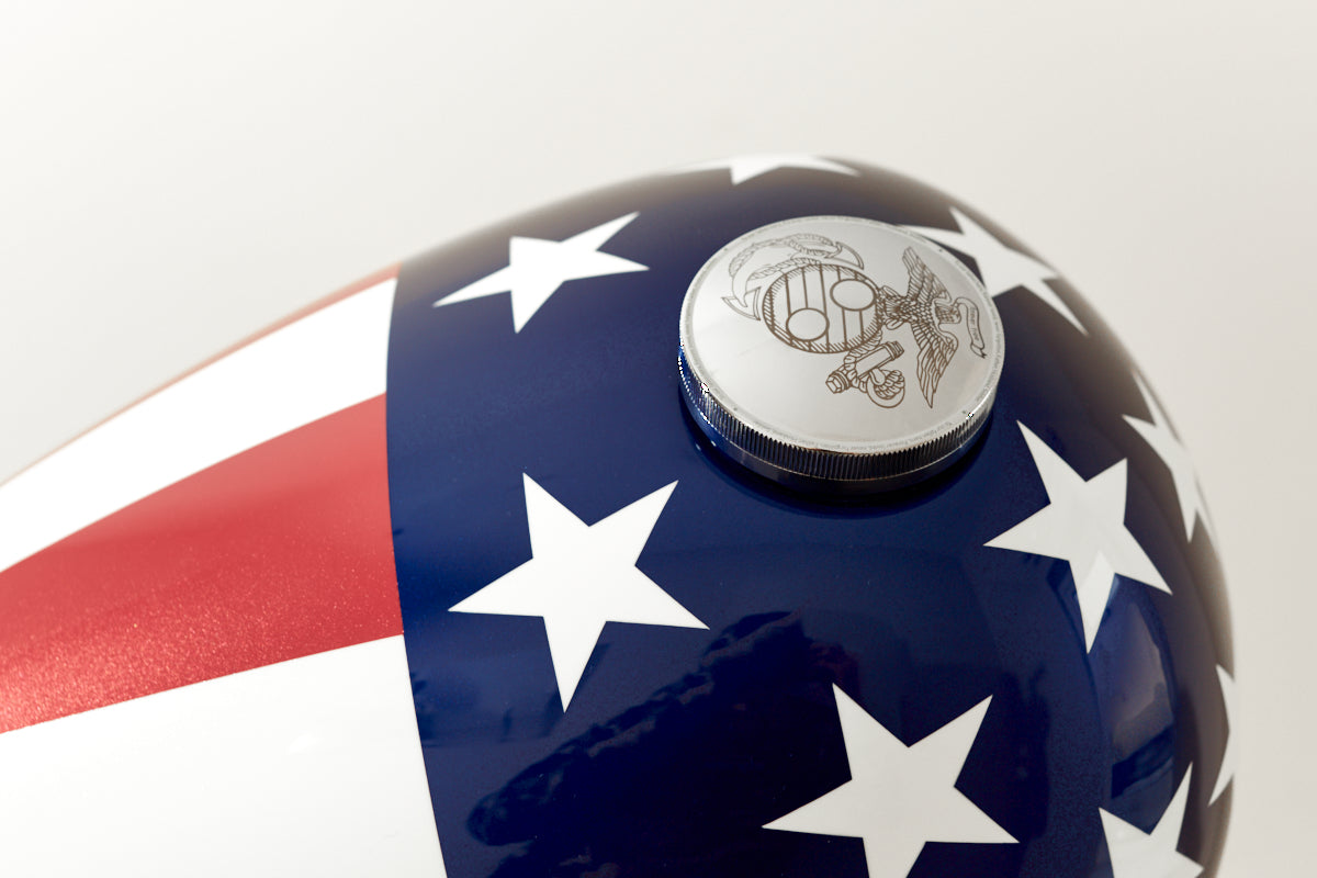 Close-up of the top of the Haus of Urns Freedom Urn, a handcrafted motorcycle gas tank cremation urn finished in high-gloss automotive lacquer with a bold American flag design in deep navy blue, red, and white stars. The chrome screw-top lid features a custom laser-engraved emblem inspired by the United States Marine Corps Eagle Globe and Anchor, sitting flush against the glossy patriotic paintwork.